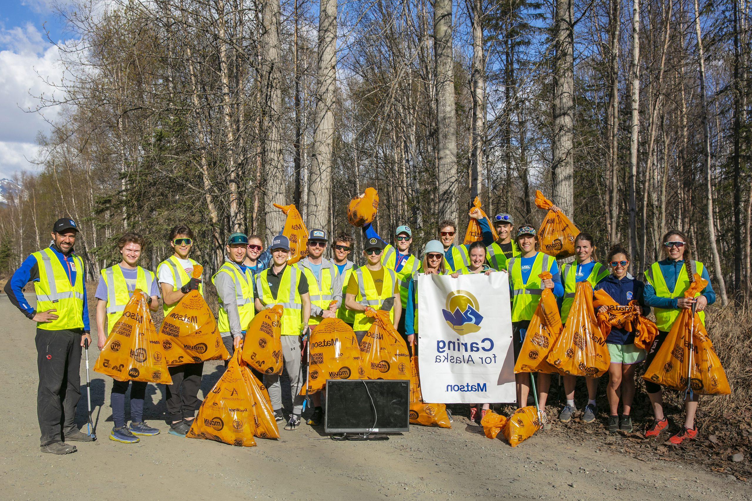 Volunteers wearing yellow safety vests pose for a group picture with their full orange trash bags and Caring For Alaska banner.